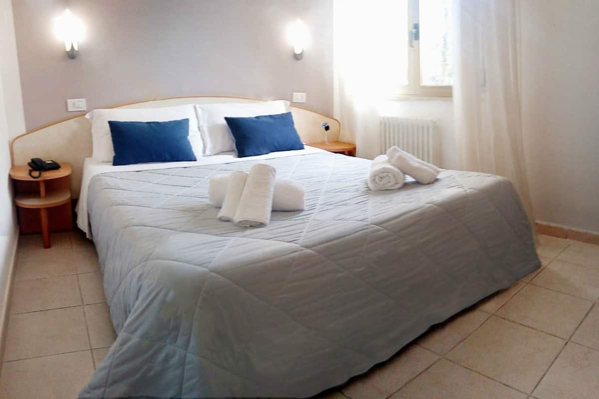 Una cama bien hecha con almohadas azules y blancas, una mesita de noche con un teléfono y una ventana con cortinas.