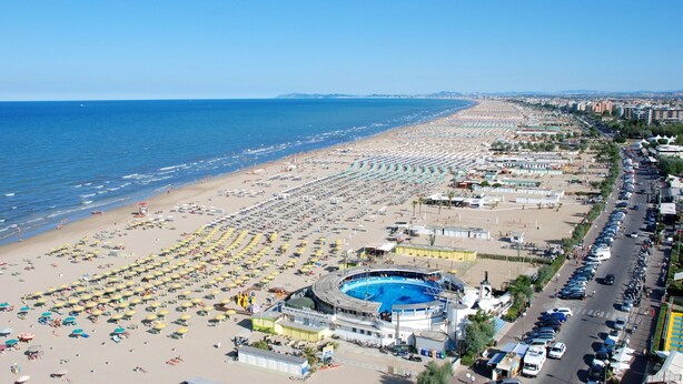 Un paisaje urbano costero con una playa extensa, una piscina y un aparcamiento.