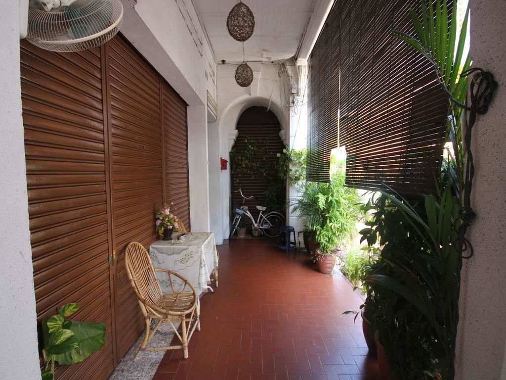 A covered outdoor area with a wooden door, a wicker chair, a table with a vase, and potted plants.