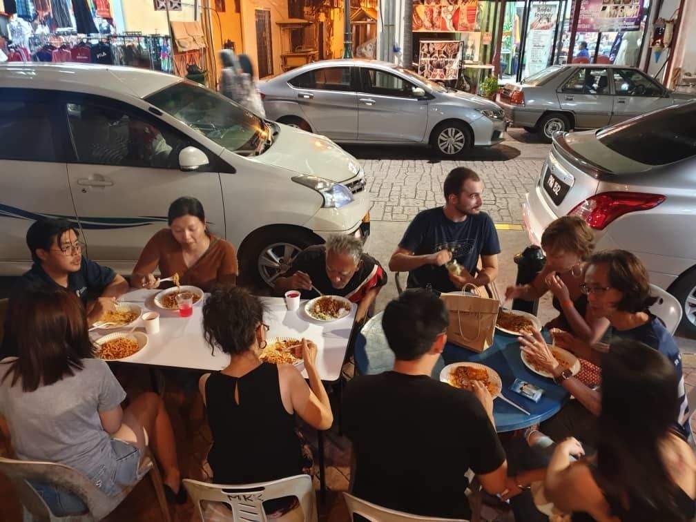 A group of people dining outdoors at a restaurant with cars parked nearby.