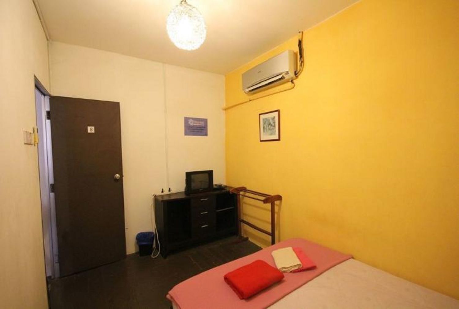 A small, compact room with a bed, a desk, and a chair. There is a door on the left and an air conditioning unit on the wall.
