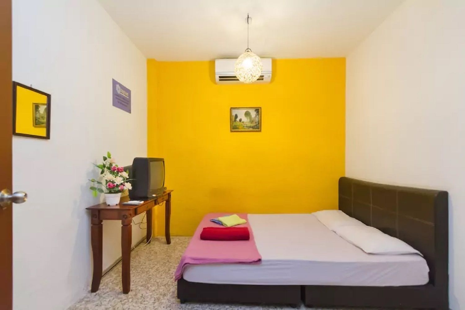 A room with a bed, a small table with a television, a flower arrangement, and a picture on the wall.