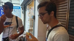 Straßenszene mit einem Mann, der an einem Tisch isst, einem Mann, der neben ihm steht, und einem geschlossenen Geschäft mit heruntergelassenen Rollläden.