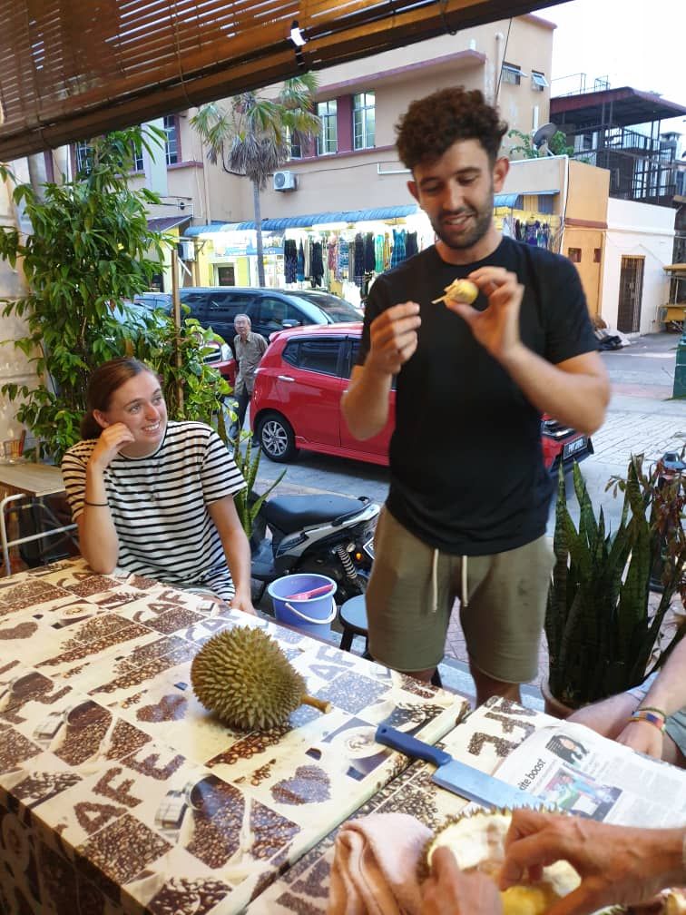 A man eating at an outdoor table with a durian fruit and a newspaper.