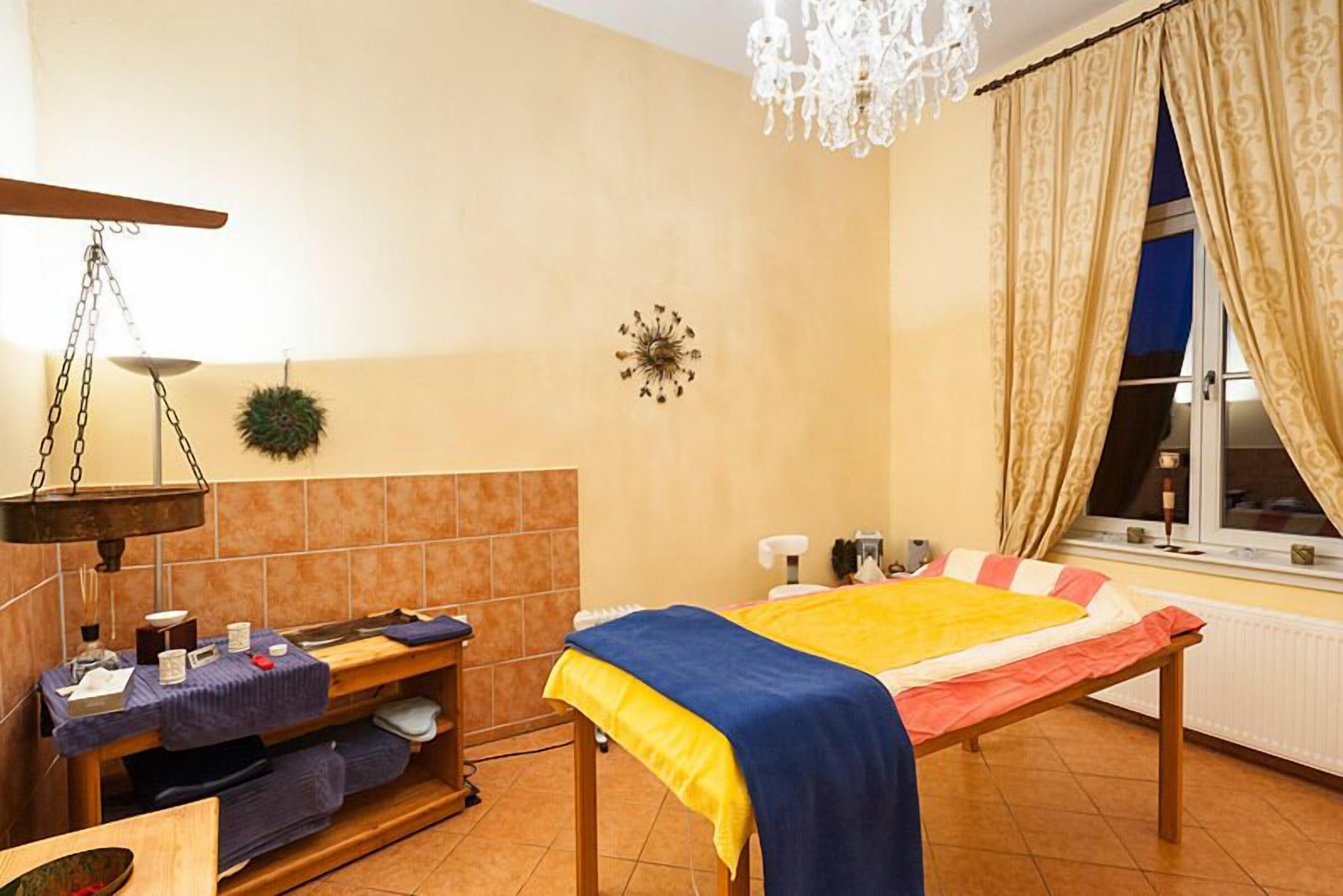 A room with a bed, a massage table, a hanging scale, a clock, and a window with curtains.