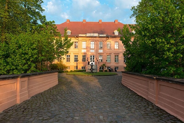 Ein Kopfsteinpflasterweg führt zu einem klassizistischen Gebäude mit rotem Dach, umgeben von üppiger Vegetation.