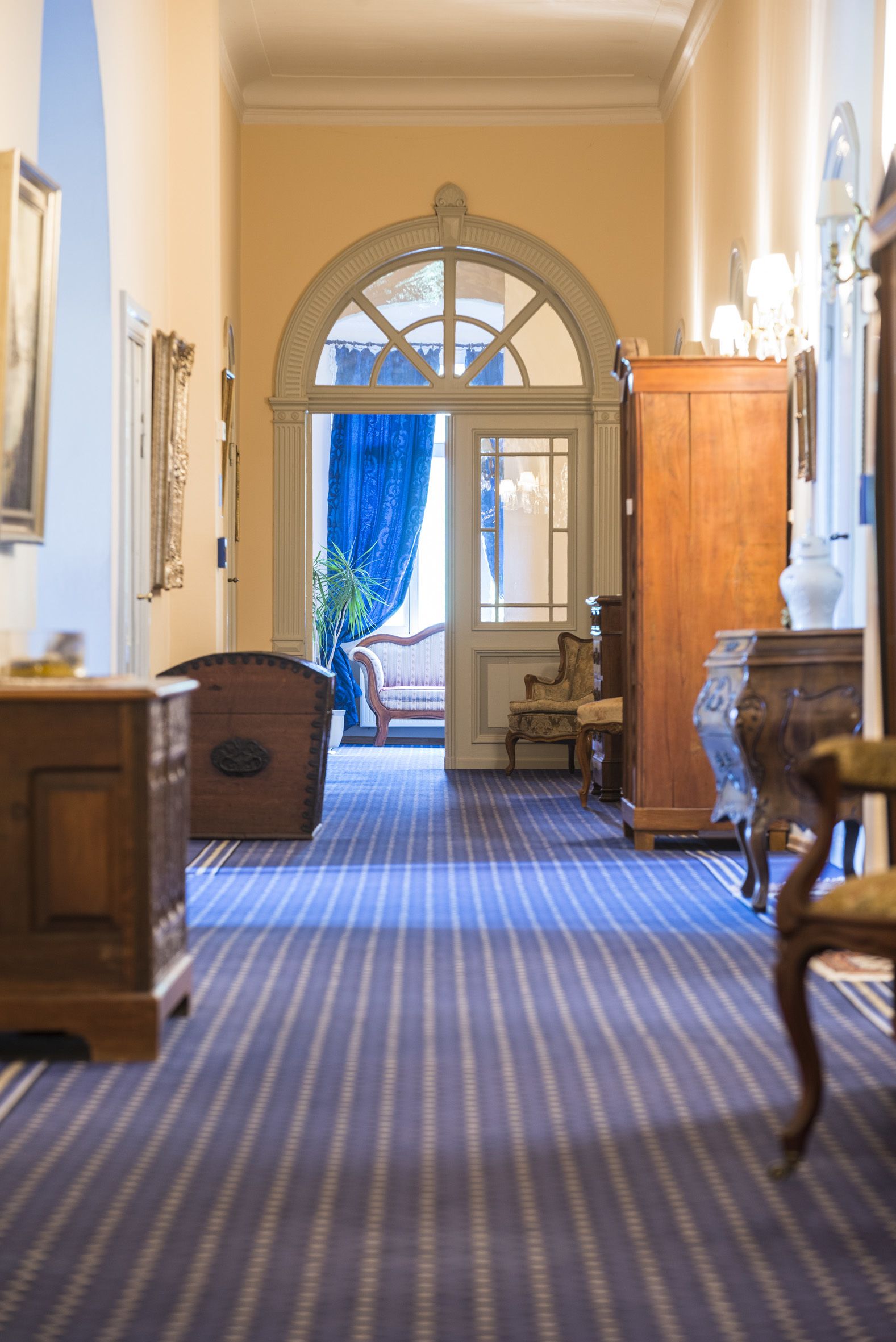Um corredor com carpete azul, mobiliário de madeira e uma grande janela em arco com cortinas azuis.