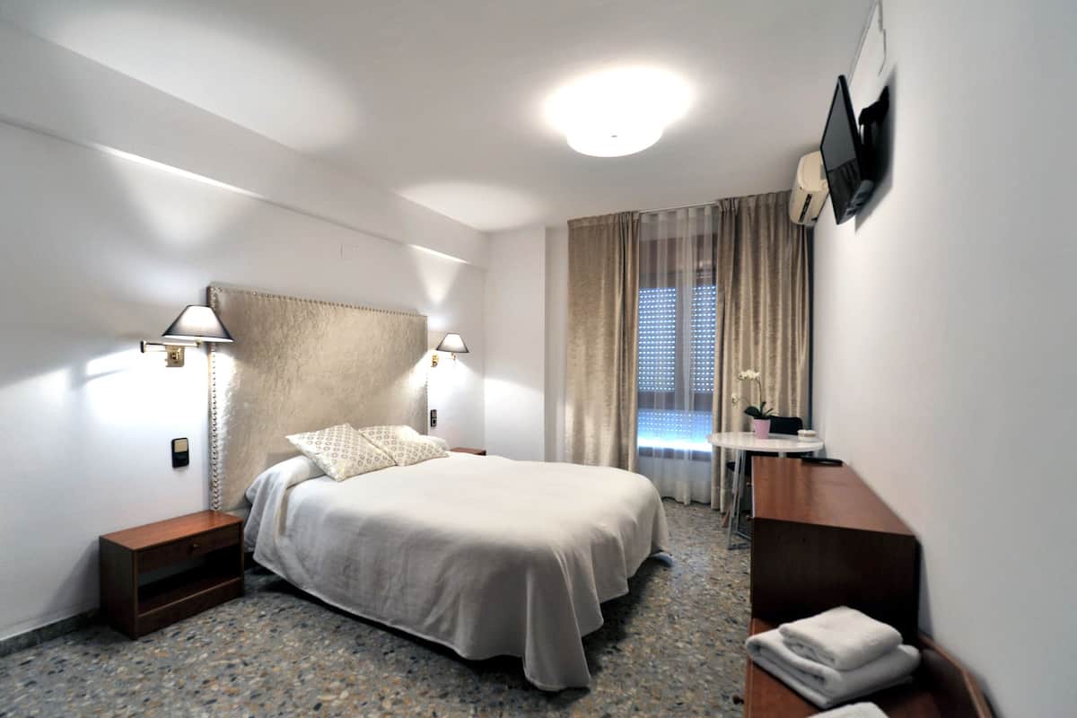 A hotel room with a bed, bedside lamps, a desk with a chair, a TV mounted on the wall, and a window with curtains.