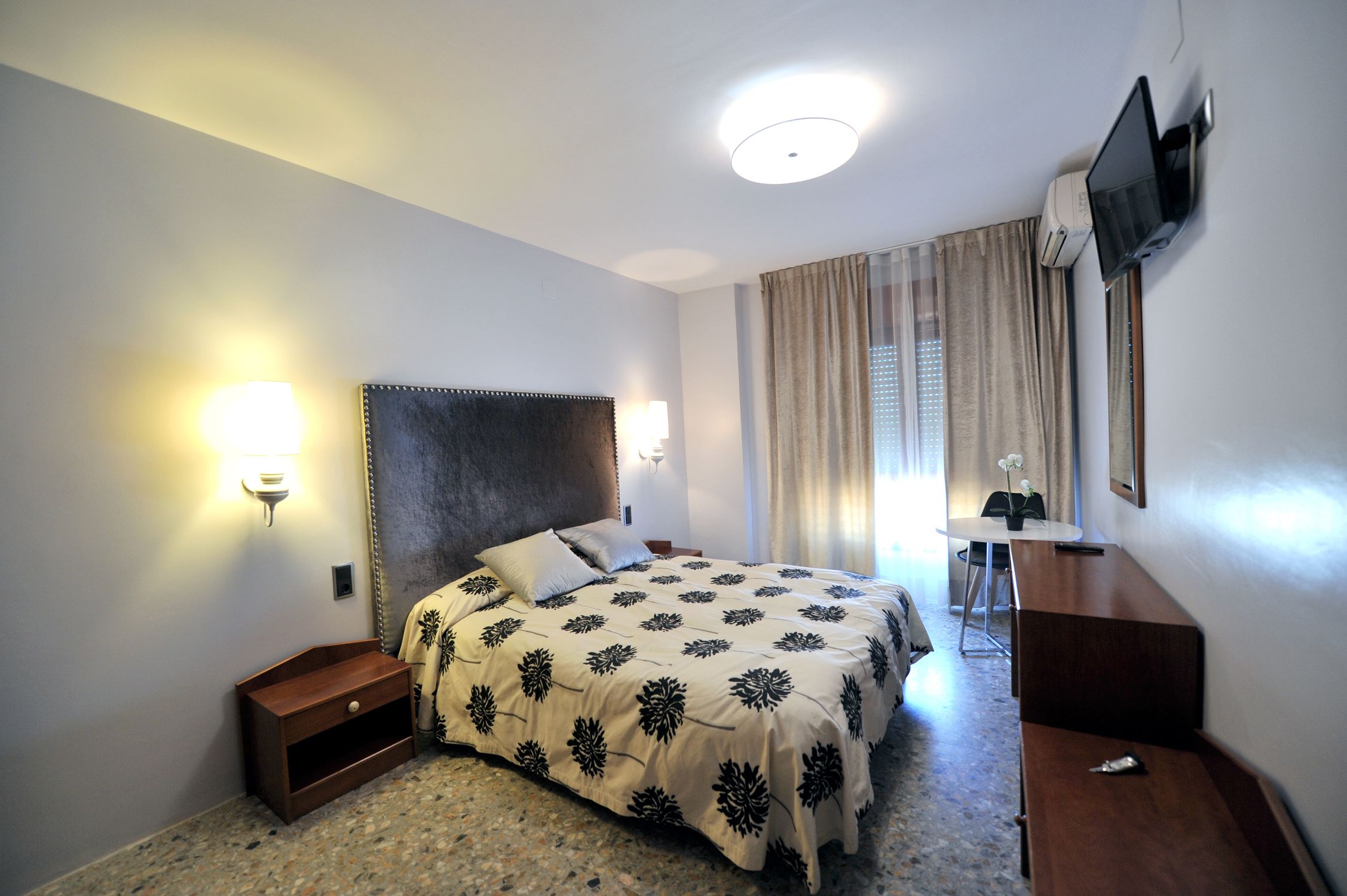 A hotel room with a bed, bedside table, lamp, TV, and a desk with a chair.