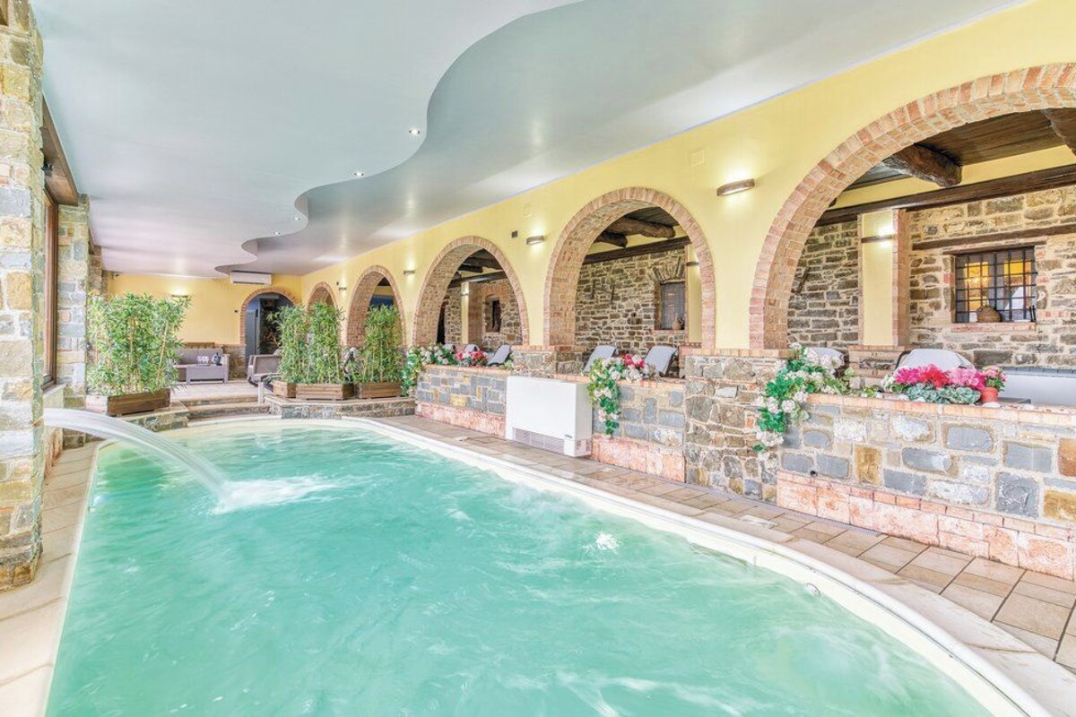 An indoor swimming pool with stone walls and arches, surrounded by potted plants and flowers.