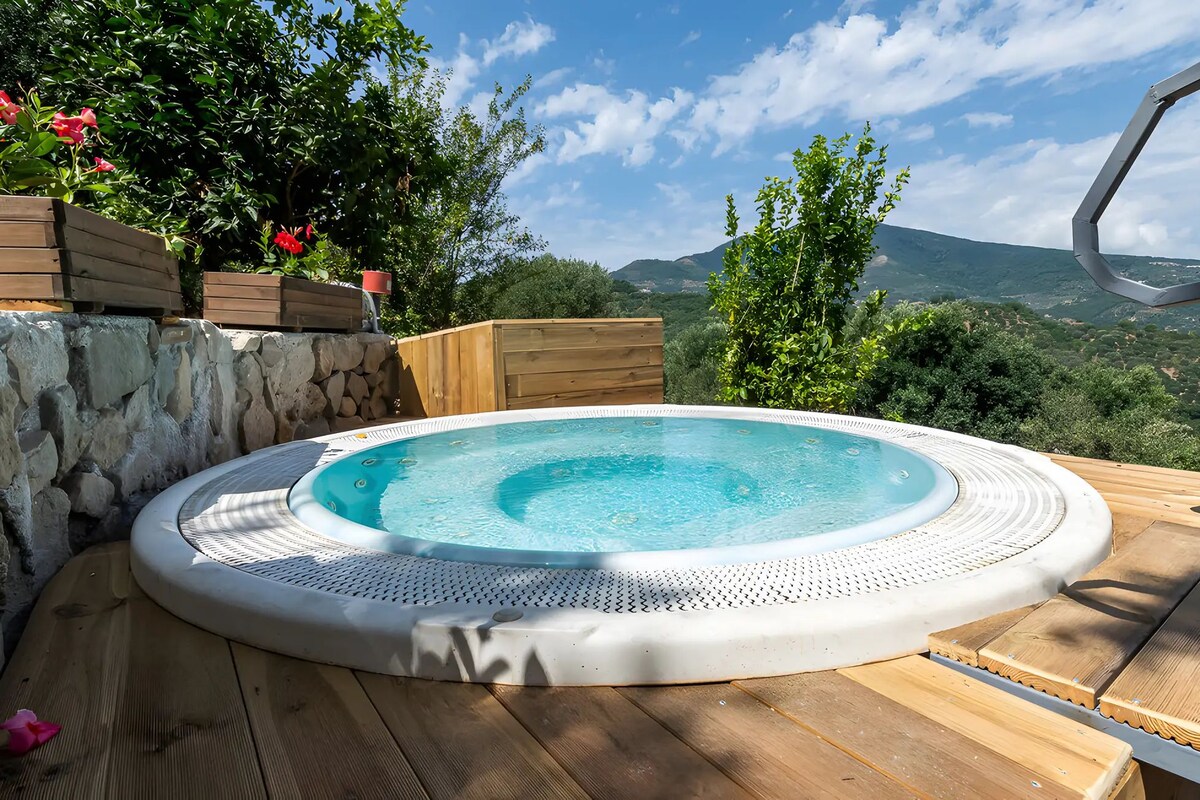 A hot tub surrounded by wooden deck, stone wall, and lush greenery with a mountainous backdrop.