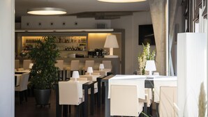 A modern restaurant with white tables and chairs, a bar area in the background, and pendant lighting.