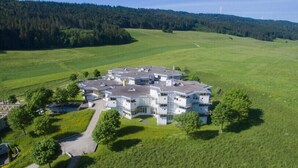 A large, modern building with multiple wings, surrounded by expansive green fields and a forested hill in the background.