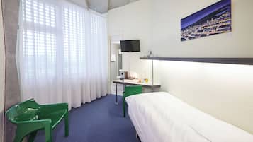 Ein Hotelzimmer mit einem Bett, einem Schreibtisch mit Stuhl, einem an der Wand befestigten Fernseher und einem Fenster mit Vorhängen.