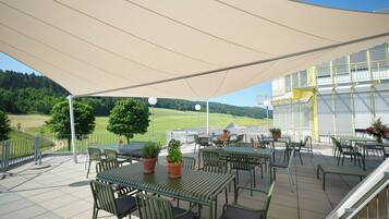 Eine Terrasse im Freien mit Tischen und Stühlen, einem großen Sonnensegel und Blick auf eine grüne Landschaft.