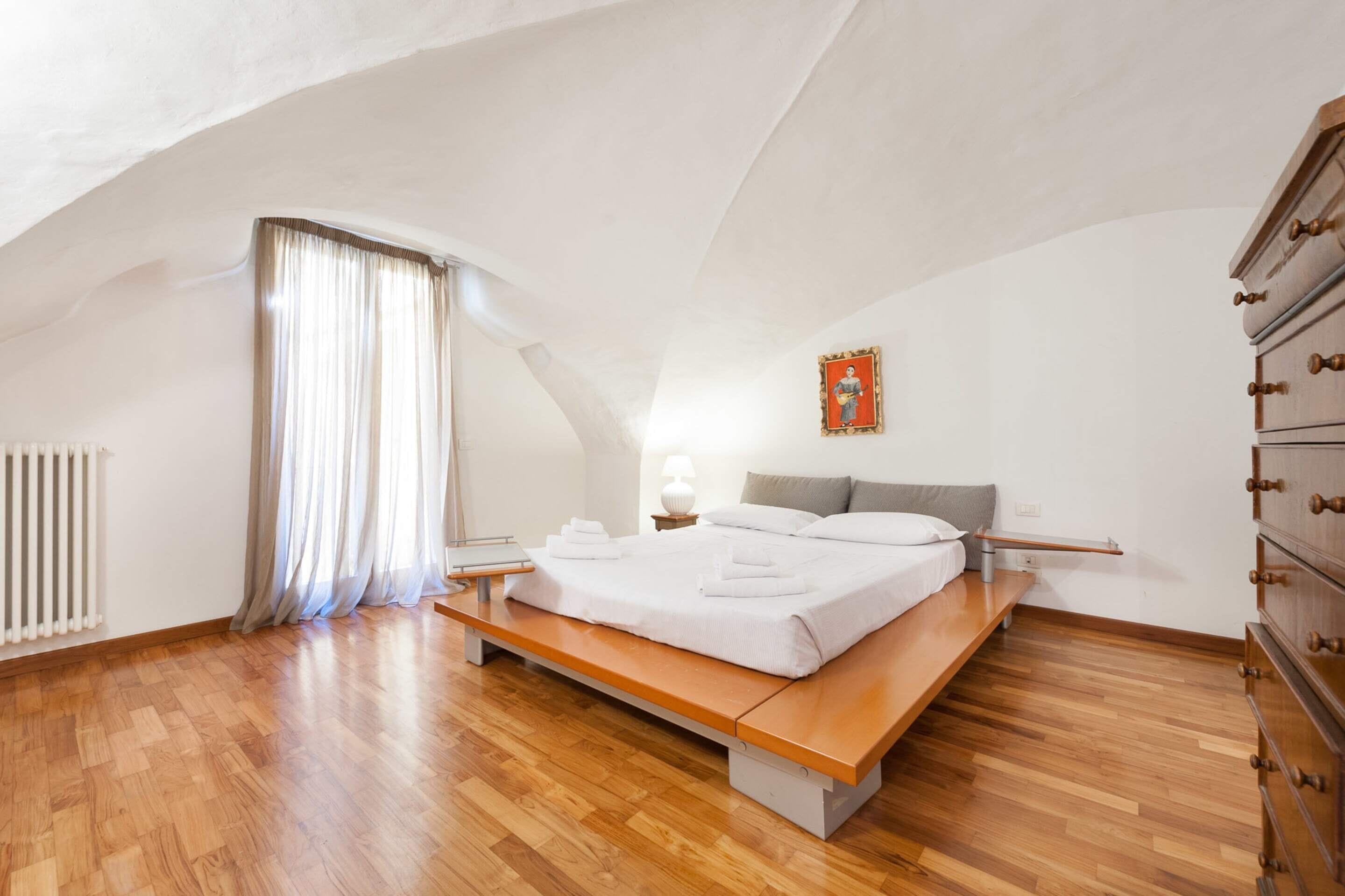 Een slaapkamer met een houten bed, een bankje, een nachtkastje, een dressoir, een radiator en een raam met gordijnen.