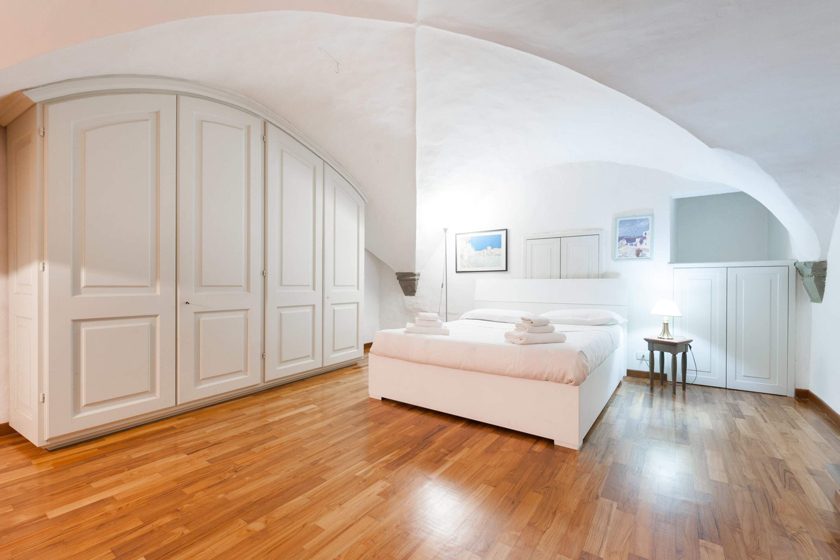 A bedroom with a wooden floor, a bed with pillows, a nightstand, and a wardrobe.