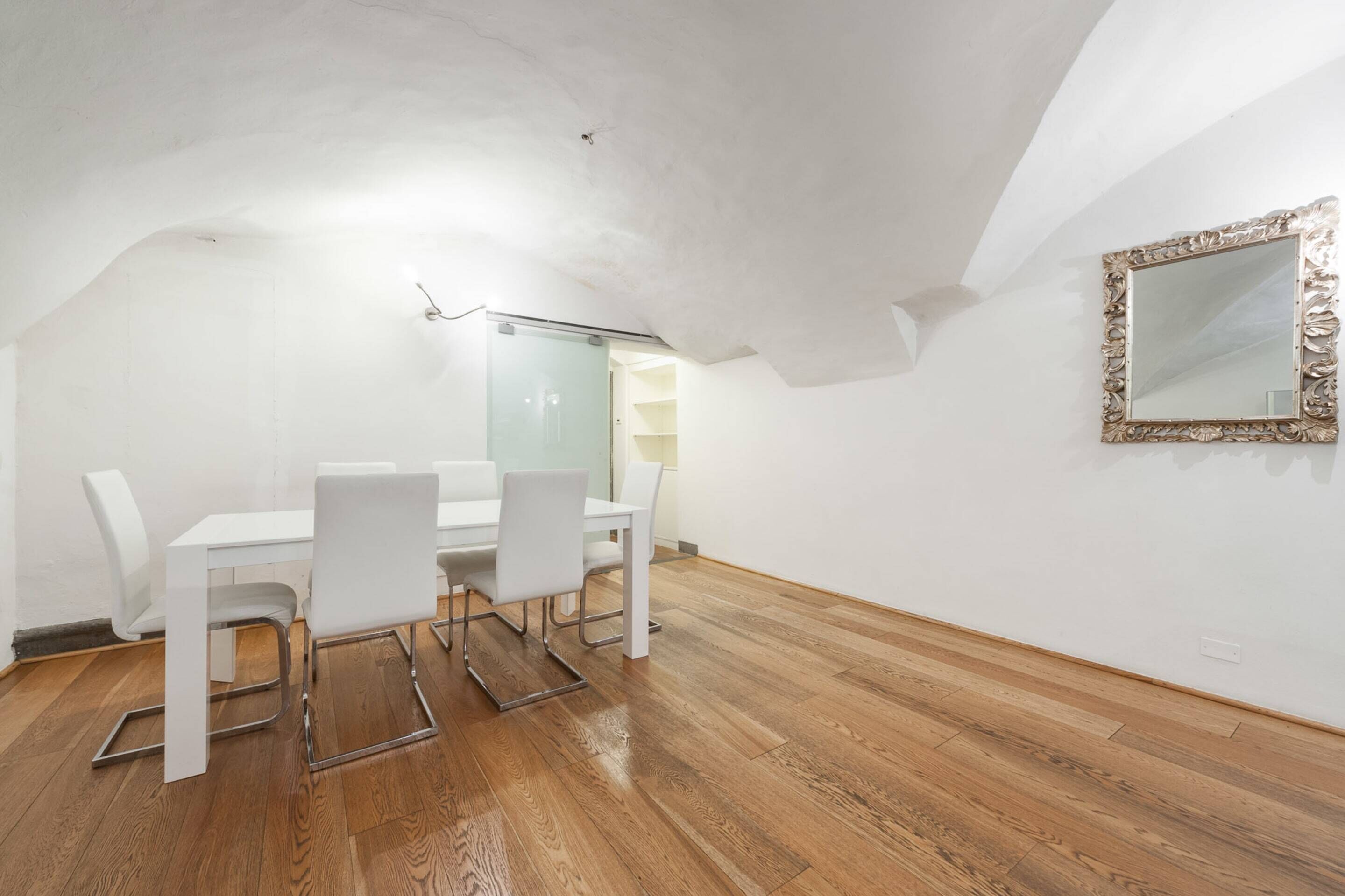 Een moderne, witte kamer met houten vloer, een witte eettafel met stoelen, en een spiegel met een versierde lijst.