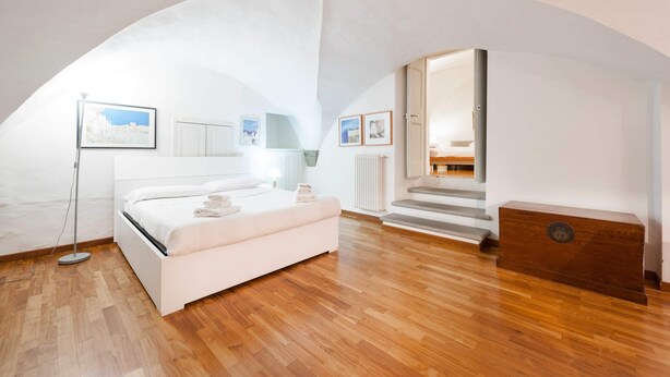 A bedroom with a white bed, wooden flooring, a standing lamp, and a view of a staircase leading to another room. - Short-let Florence Pontevecchio Suite (Florence)