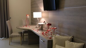A modern hotel room with a dining area featuring a table set with candles, glasses, and a lamp. There is a TV mounted on the wall and a sofa with cushions. - Hotel Delle Canne (Amantea)