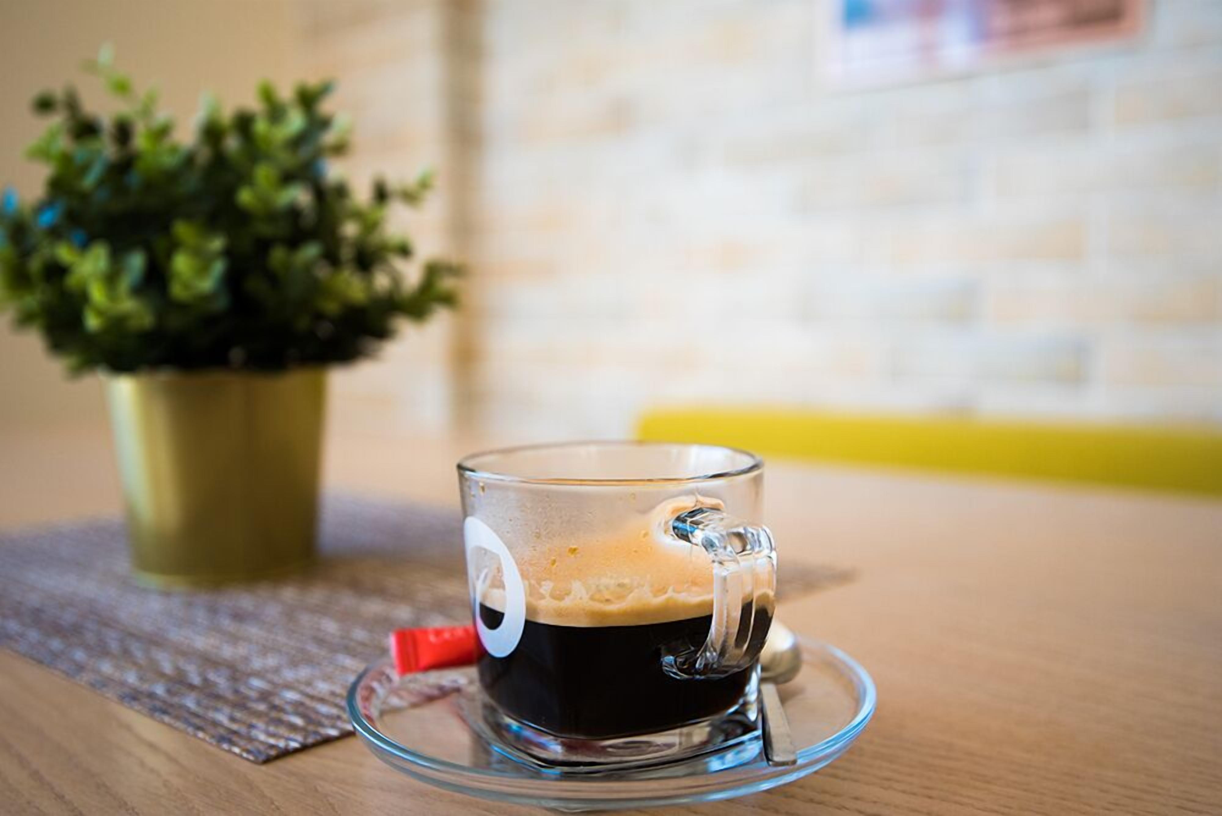 A clear glass cup of coffee on a saucer with a metal handle, placed on a wooden table.