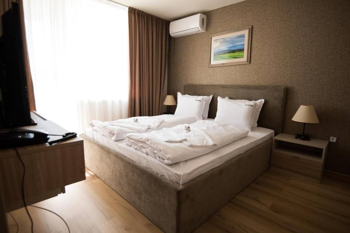 A hotel room with a bed, two bedside tables, a desk with a computer, and an air conditioning unit.