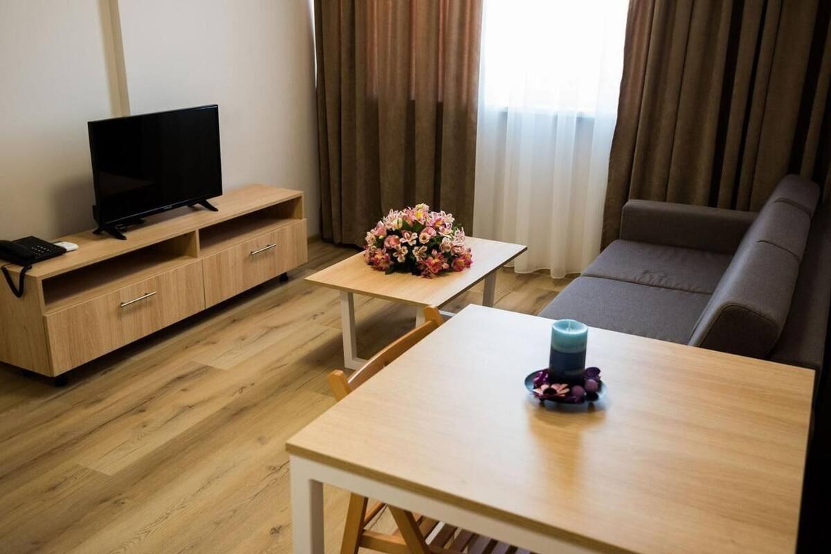 A hotel room with a TV on a wooden stand, a small table with a vase of flowers, a sofa, and a wooden floor.