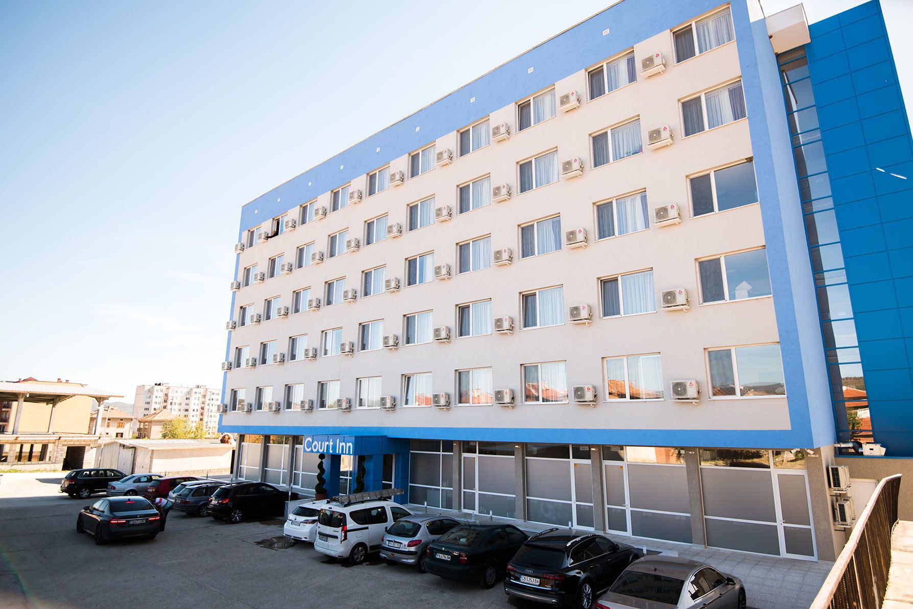 A multi-story hotel with a blue and white facade, air conditioning units, and a parking lot with several cars.