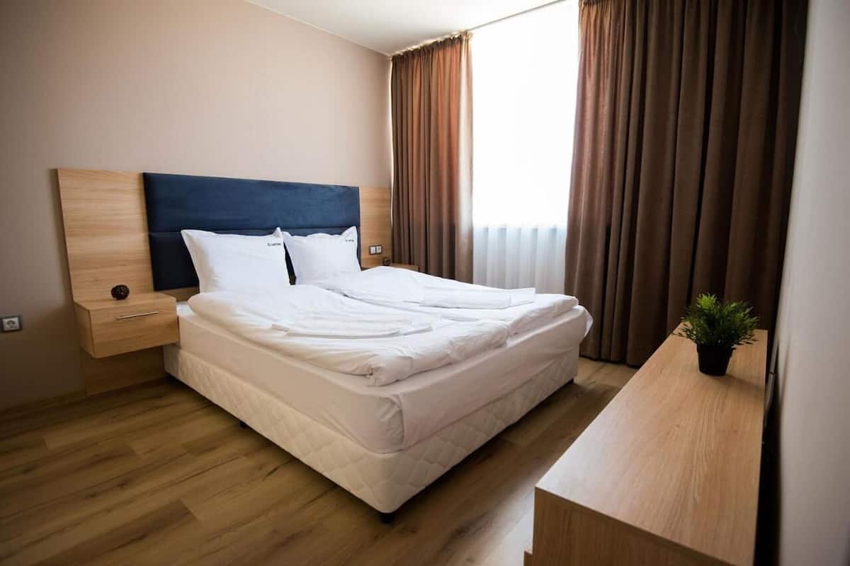 A neatly made bed in a hotel room with a wooden headboard, a bedside table with a lamp, and a potted plant on a wooden desk.