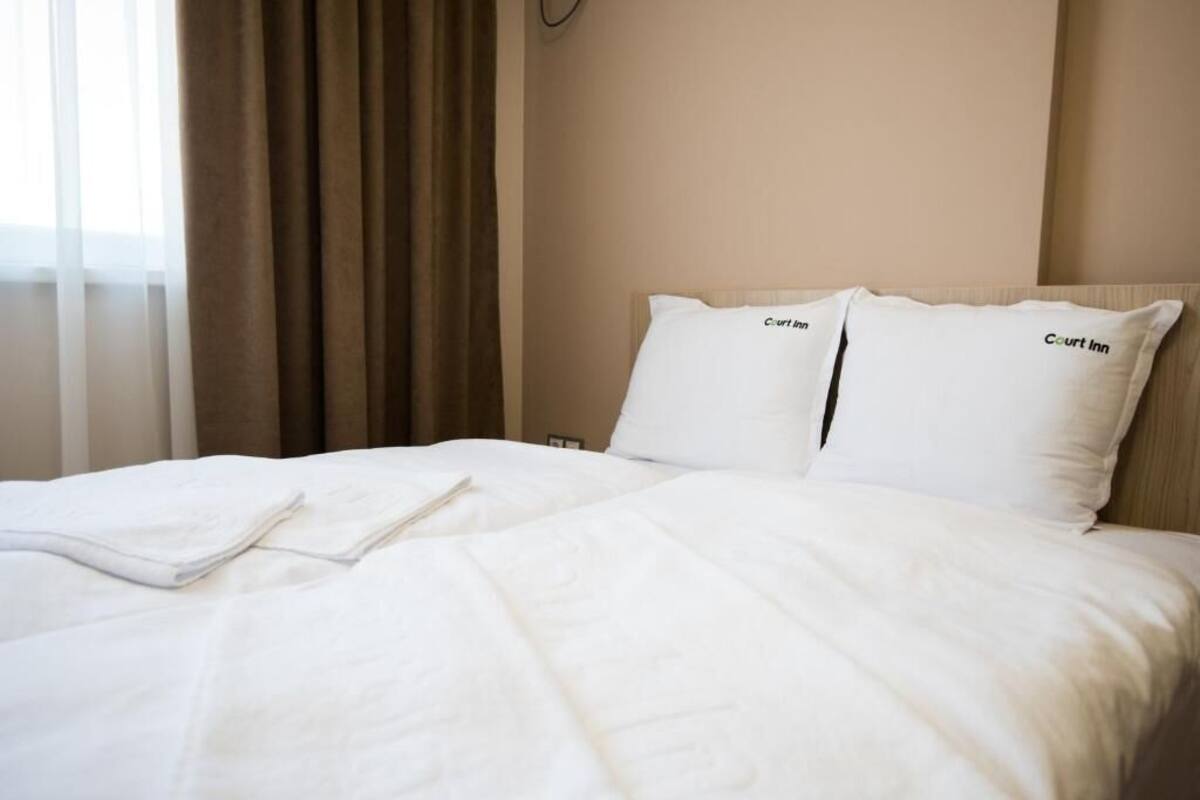 A hotel room with a neatly made bed, two pillows with the hotel name, and a window with curtains.