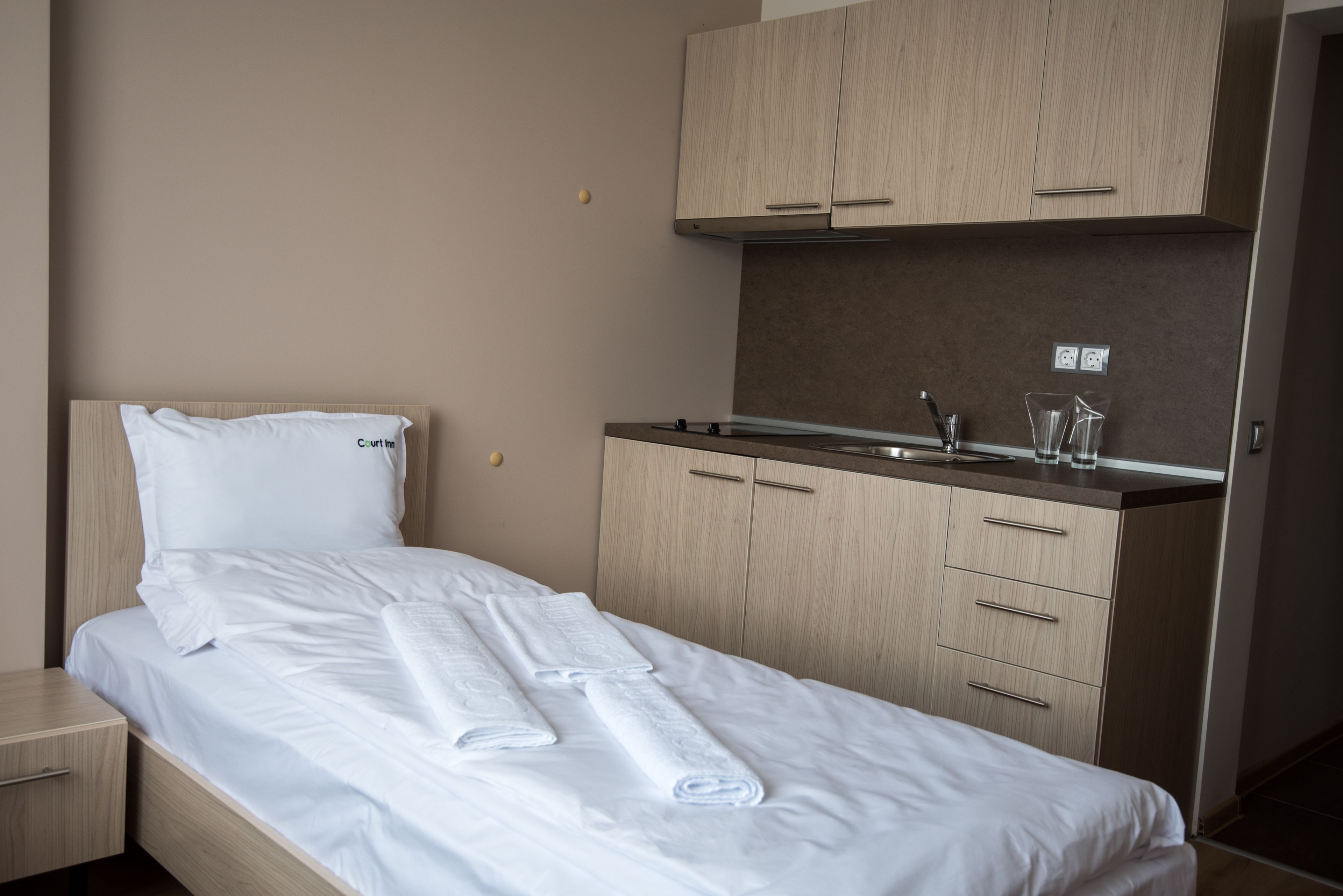 Hotelkamer met een bed, een klein nachtkastje, een ingebouwde kitchenette met een spoelbak en kastjes, en een raam.