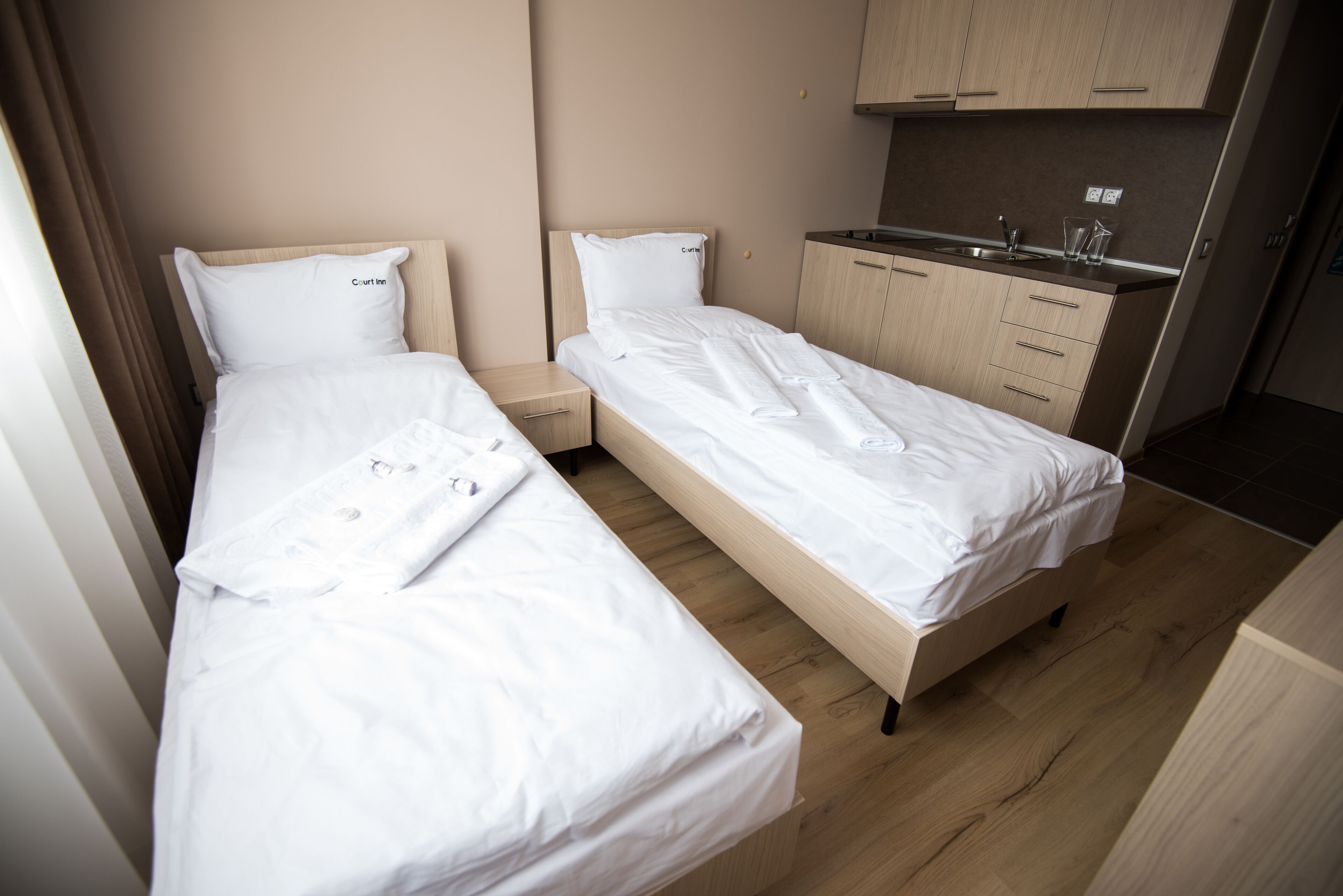 A hotel room with two single beds, a small kitchenette, and a wooden floor.