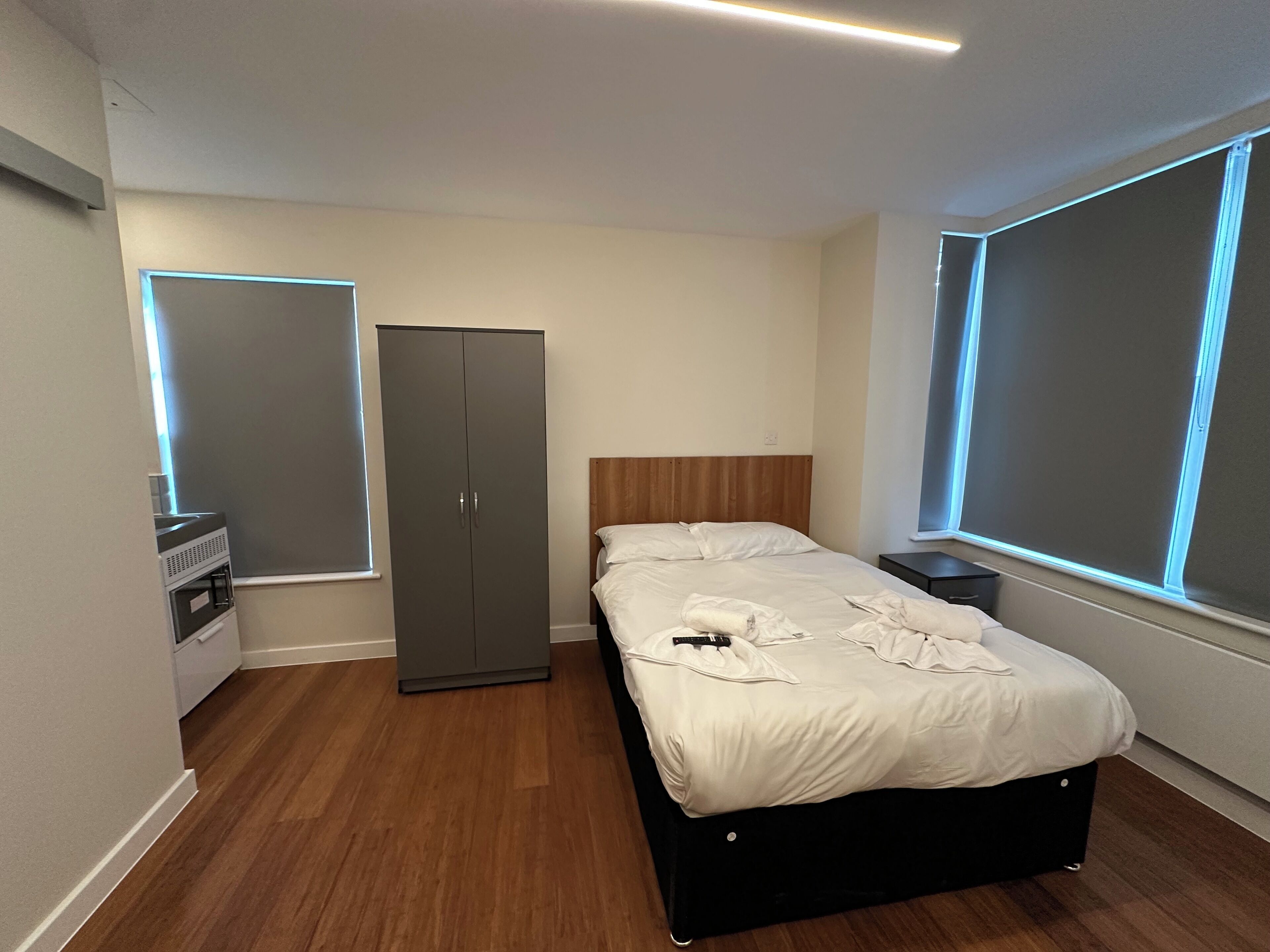 Un dormitorio con cama, armario, mesitas de noche y ventanas amplias con persianas.
