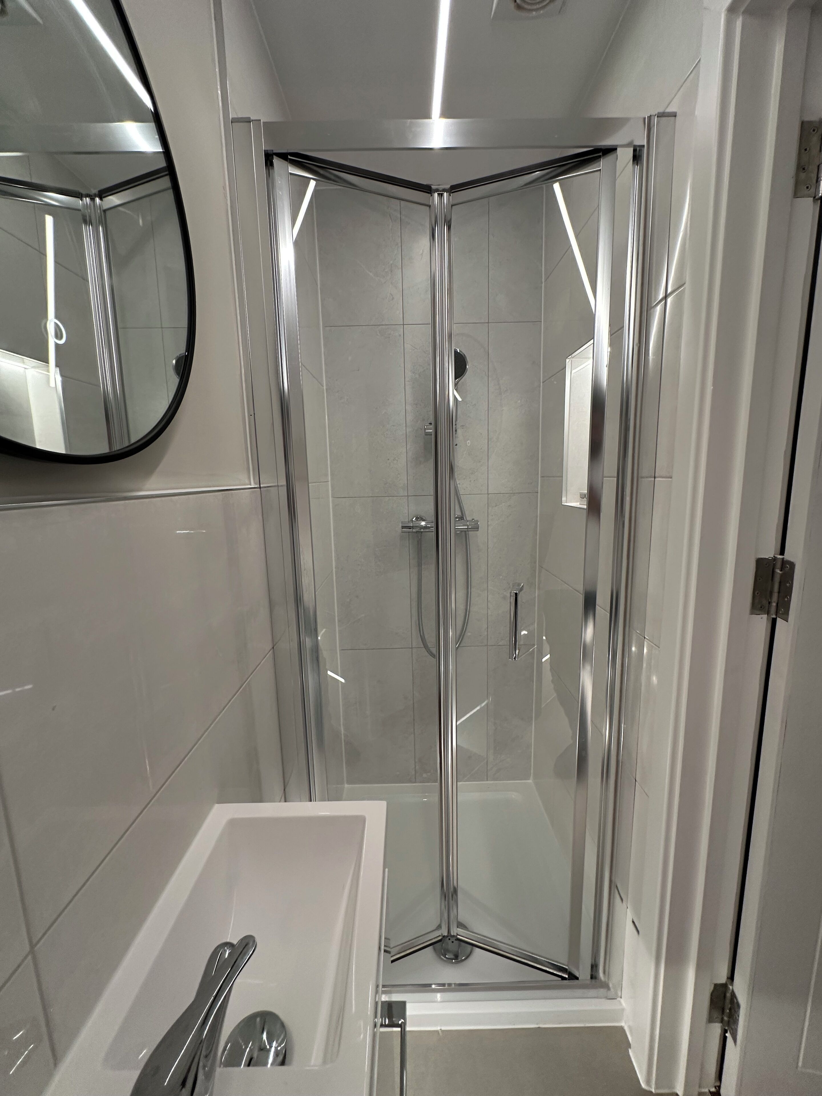 A modern bathroom with a glass-enclosed shower, a white bathtub, and a round mirror on the wall.