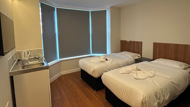 A modern hotel room with two beds, a small kitchenette, and a corner window with built-in lighting. - Contractors Guest House (High Wycombe)