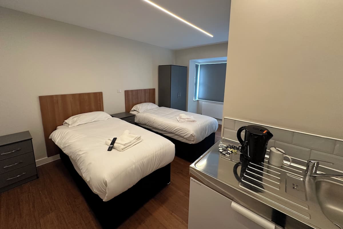 Una camera d'albergo con due letti, un angolo cottura, una macchina per il caffè e una finestra con persiane.