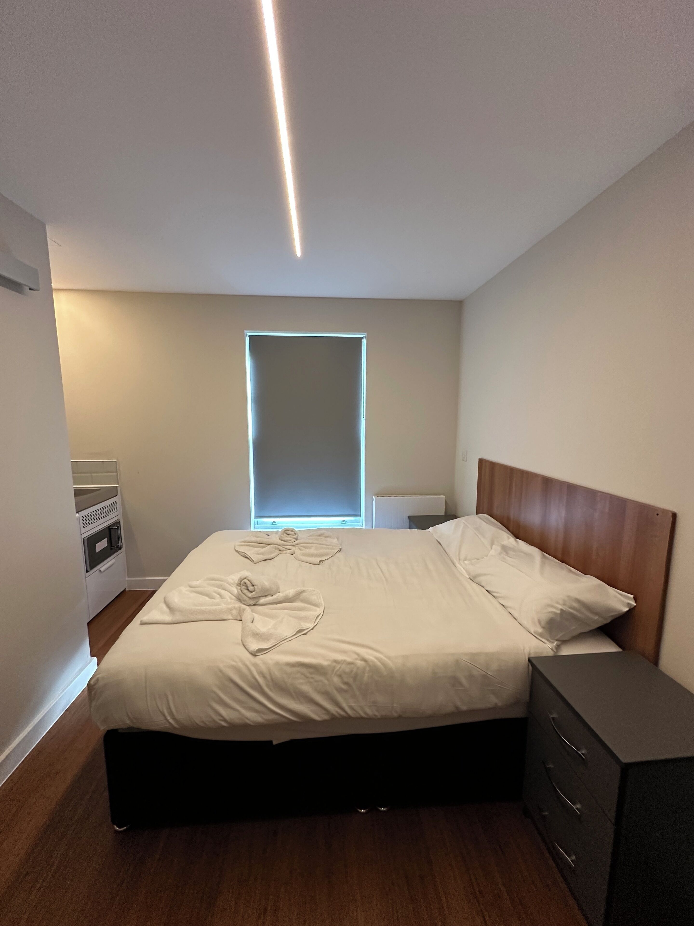 A modern bedroom with a bed, bedside tables, a dresser, a built-in oven, and a window with blinds.