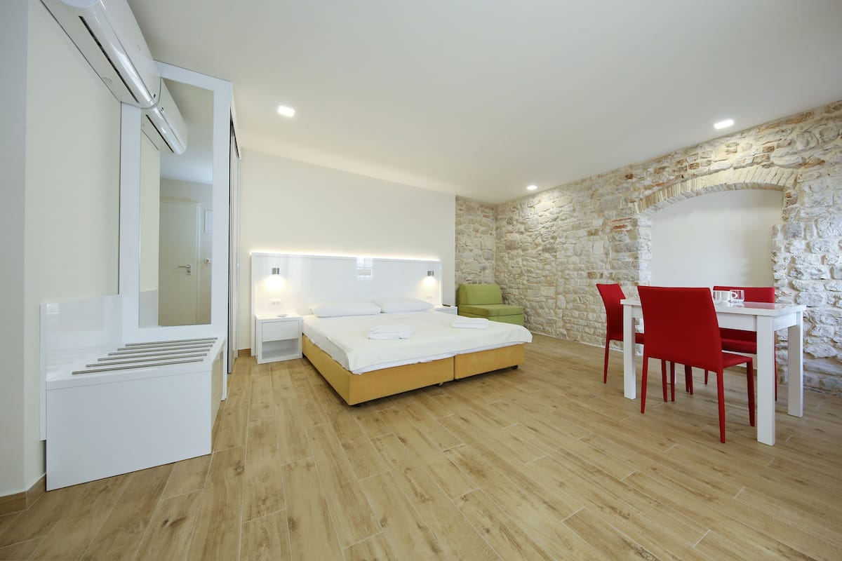 A modern bedroom with a wooden floor, a bed with white linens, a green chair, a white table with red chairs, and a stone wall.