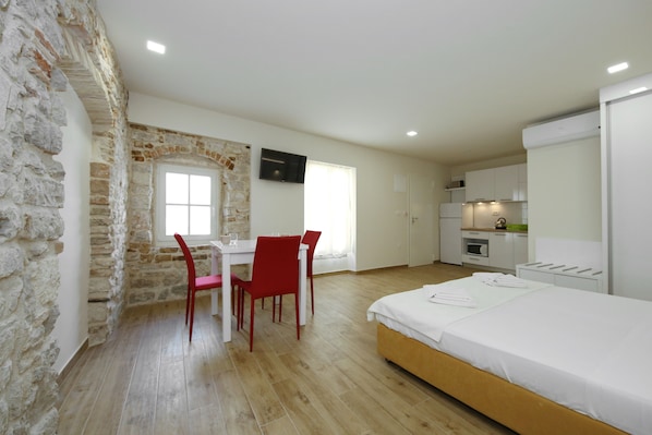 A bedroom with a wooden bed, a dining area with a table and chairs, a TV mounted on the wall, and a kitchen area with cabinets and a countertop.