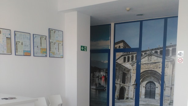 A reception area with a white desk, chairs, and a wall with framed documents. There is a door with a blue frame leading to an outdoor area with a view of a building.