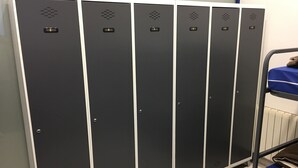 A row of black lockers with silver trim, a blue mattress on a metal frame, and a white radiator in the background.