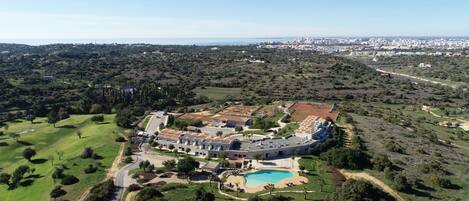 An aerial view of a large, circular building complex with a swimming pool, surrounded by green fields and trees.