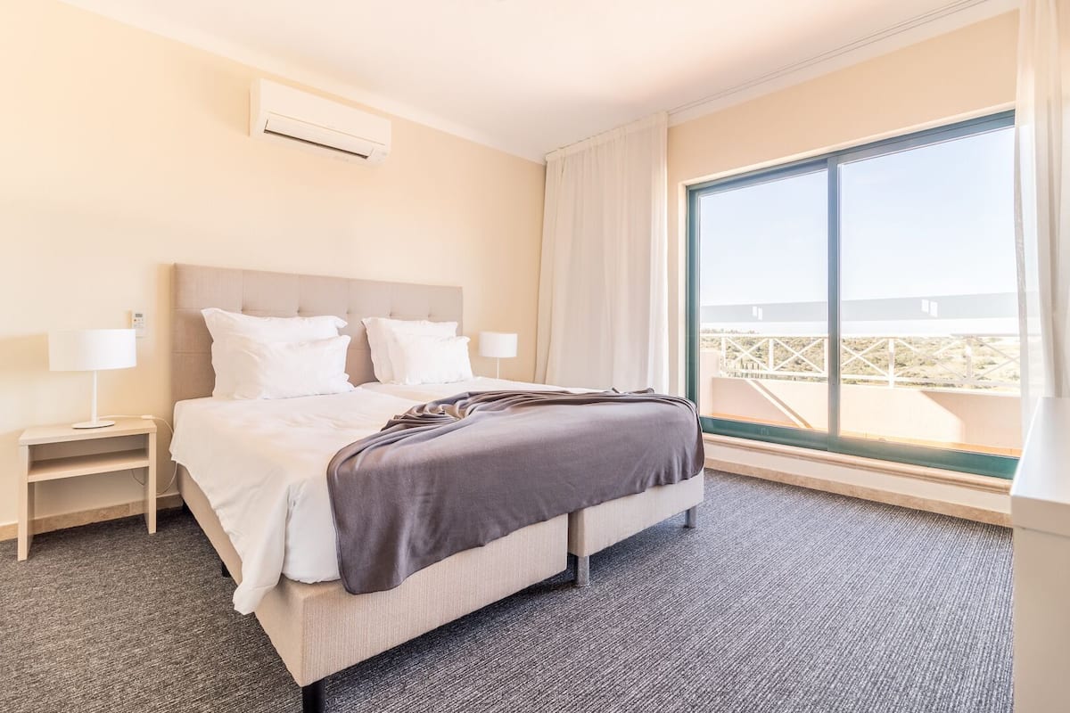 Une chambre d’hôtel comprenant un lit, des tables de chevet, un appareil de climatisation, une grande fenêtre avec vue et un balcon.