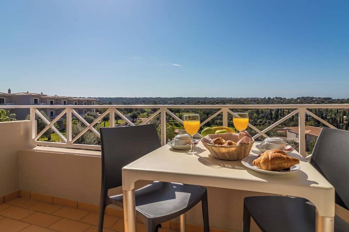 Un balcon avec une table préparée pour le petit-déjeuner, comprenant des croissants, des fruits et du jus d’orange, et offrant une vue sur un paysage magnifique.