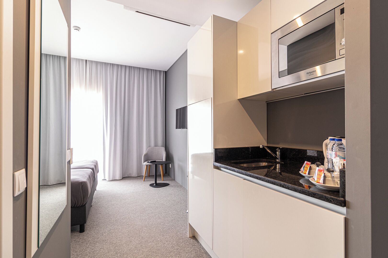 A compact hotel room with a kitchenette, a bed, a chair, and a view of the outside through a window with curtains.