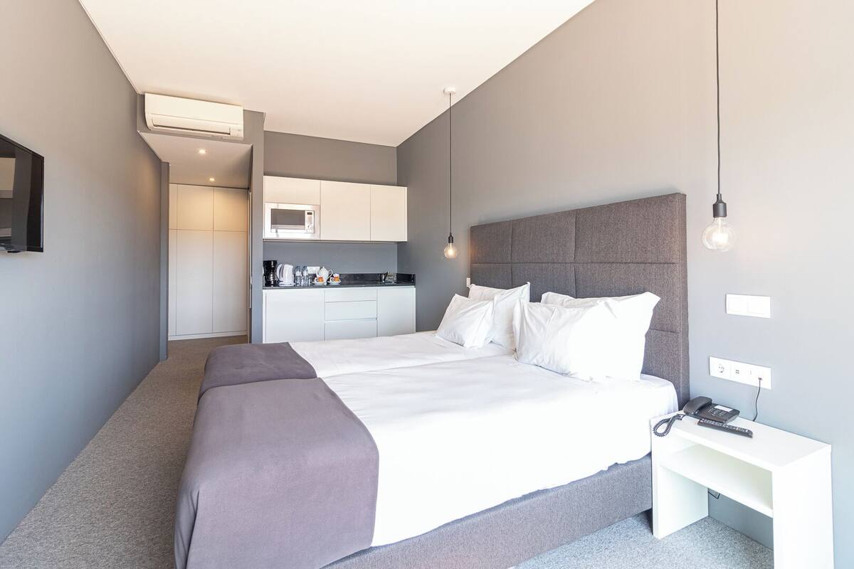 Une chambre d’hôtel moderne avec un grand lit, une table de chevet avec un téléphone, une petite kitchenette et un mur d’accent de couleur grise.
