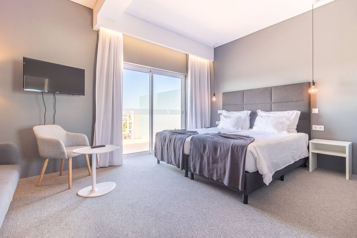 Une chambre d’hôtel moderne équipée d’un lit, d’une chaise, d’une petite table, d’une télévision et d’un balcon avec des rideaux.