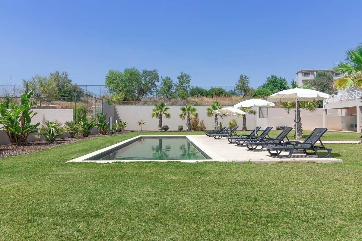 Une piscine entourée de chaises longues et de parasols, avec une pelouse bien entretenue et des palmiers.