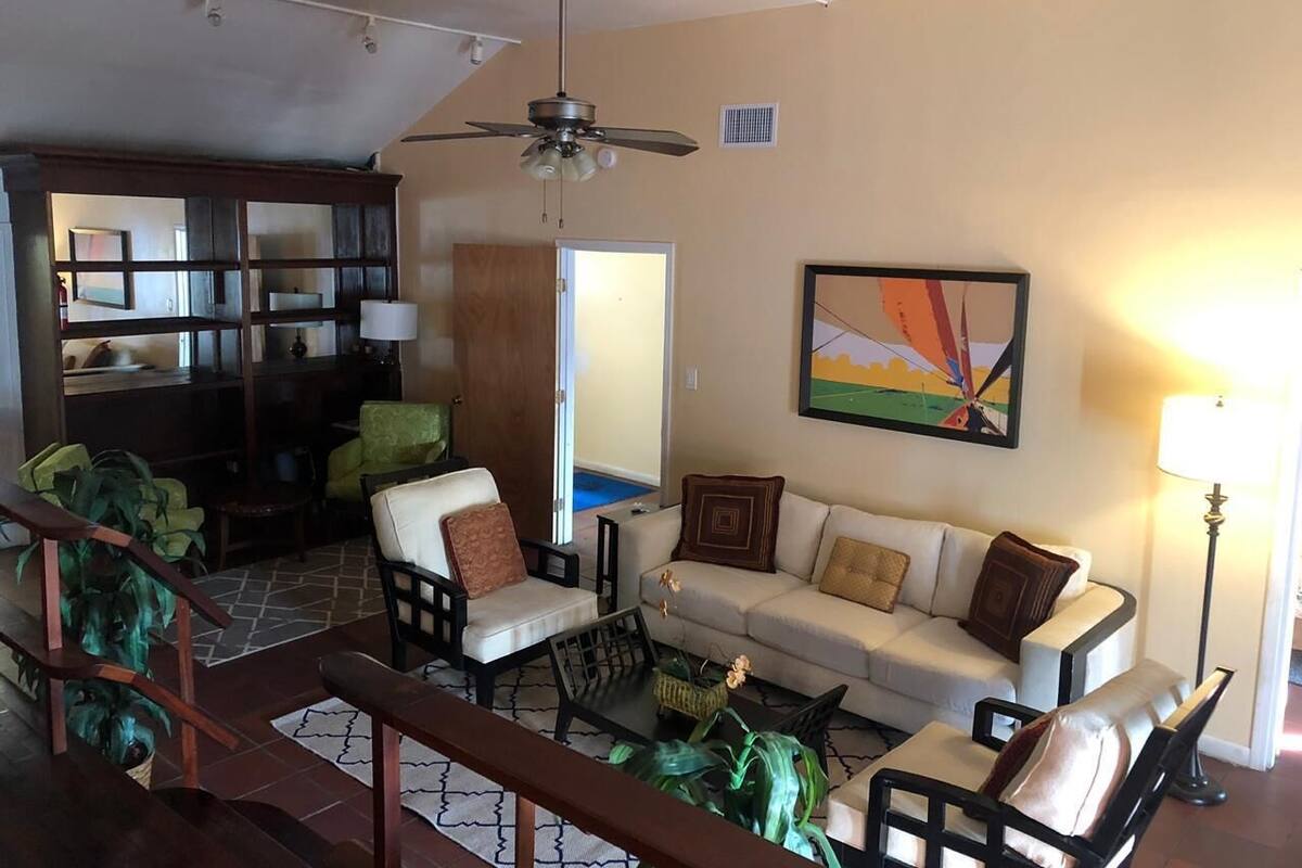 A living room with a sofa, chairs, a coffee table, a ceiling fan, a painting, and a staircase.
