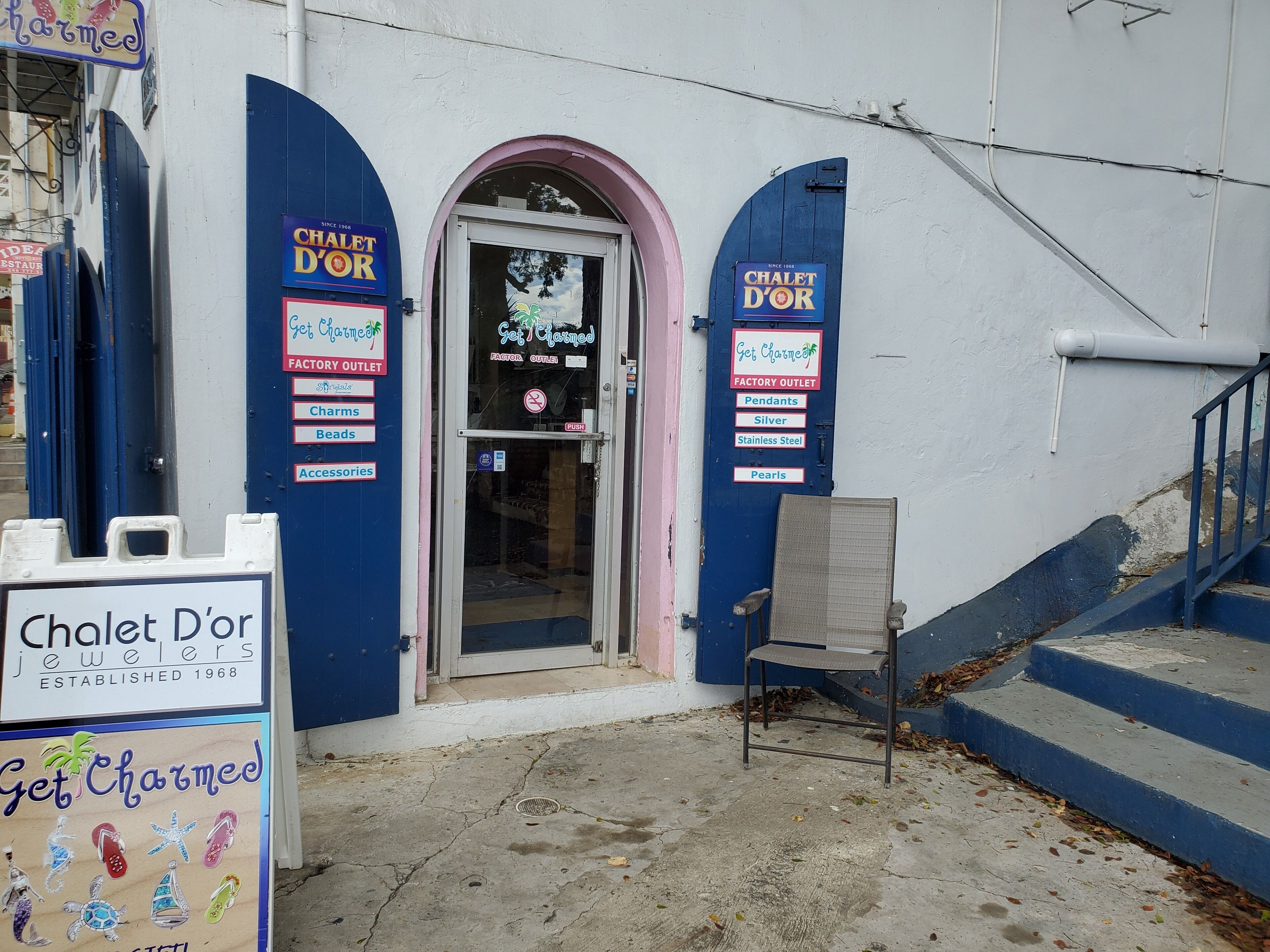 A storefront with a blue and white facade, featuring a glass door entrance and a signboard advertising jewelry items like charms, beads, and accessories.